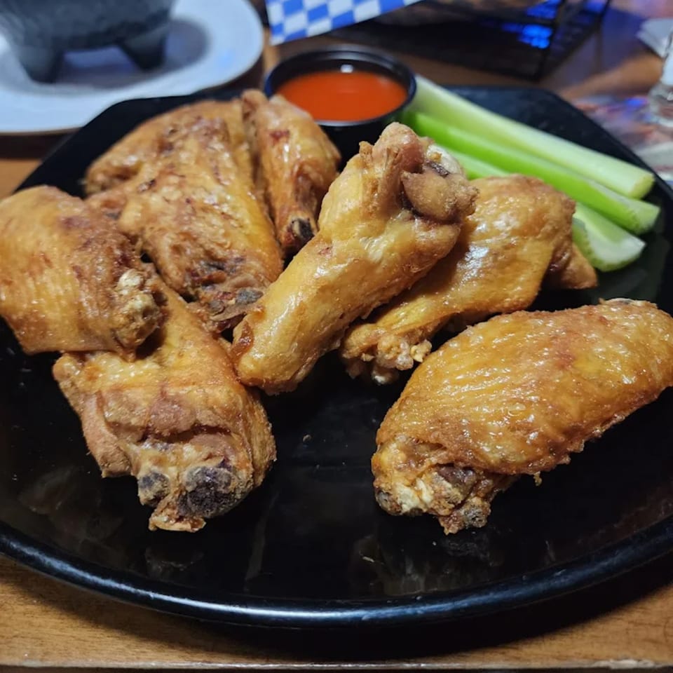 Golden-brown chicken wings with a side of hot sauce and celery sticks on a black plate.