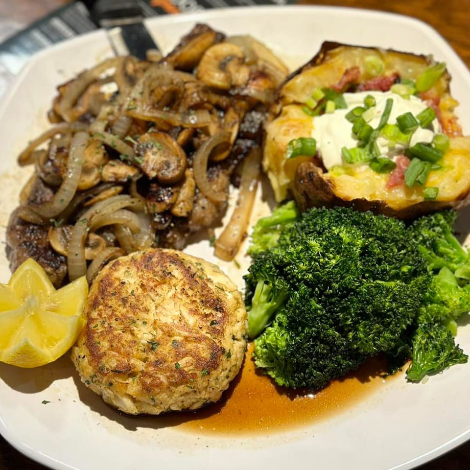 A bar-and-grill platter with steak, mushrooms, crab cake, baked potato, broccoli, and lemon.