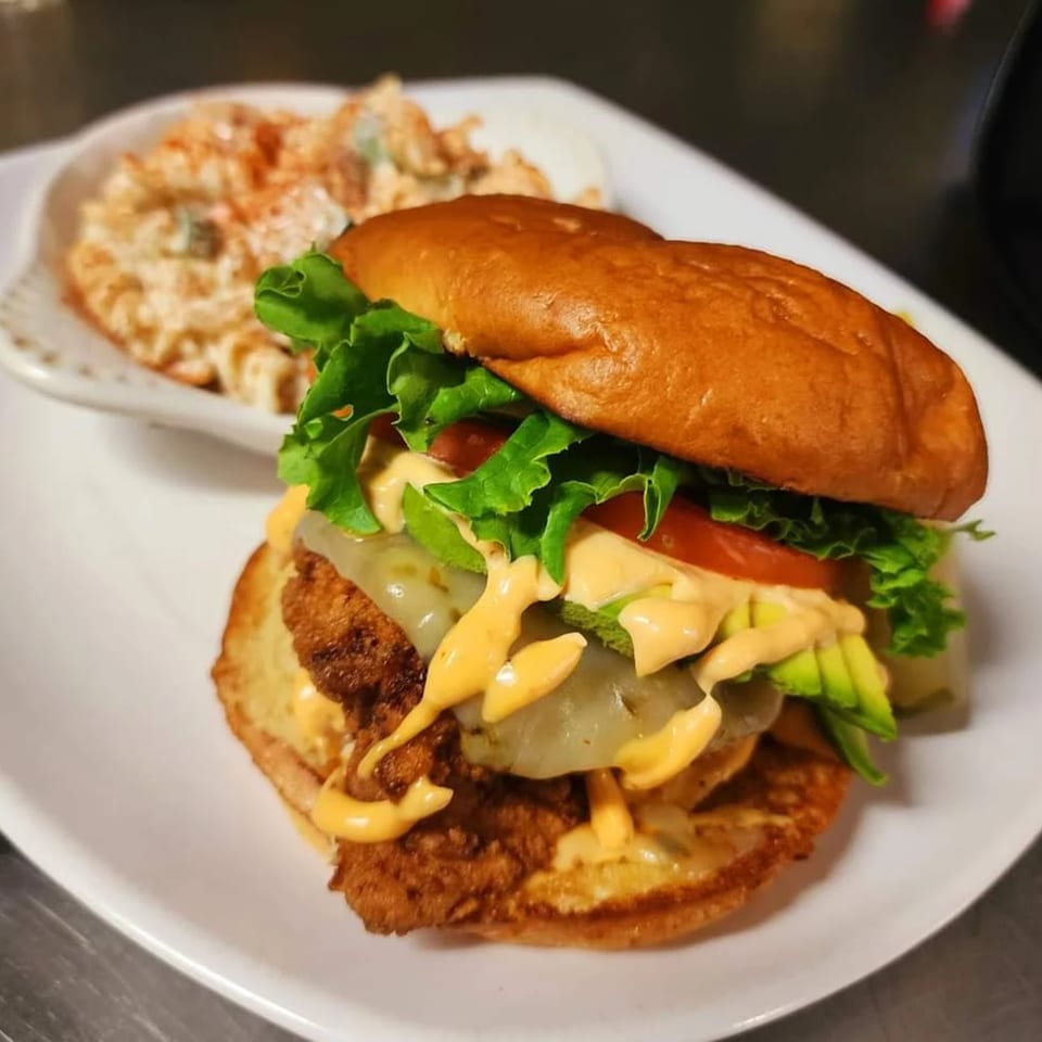 Crispy chicken sandwich with lettuce, tomato, avocado, cheese, and sauce, served with a side of potato salad.