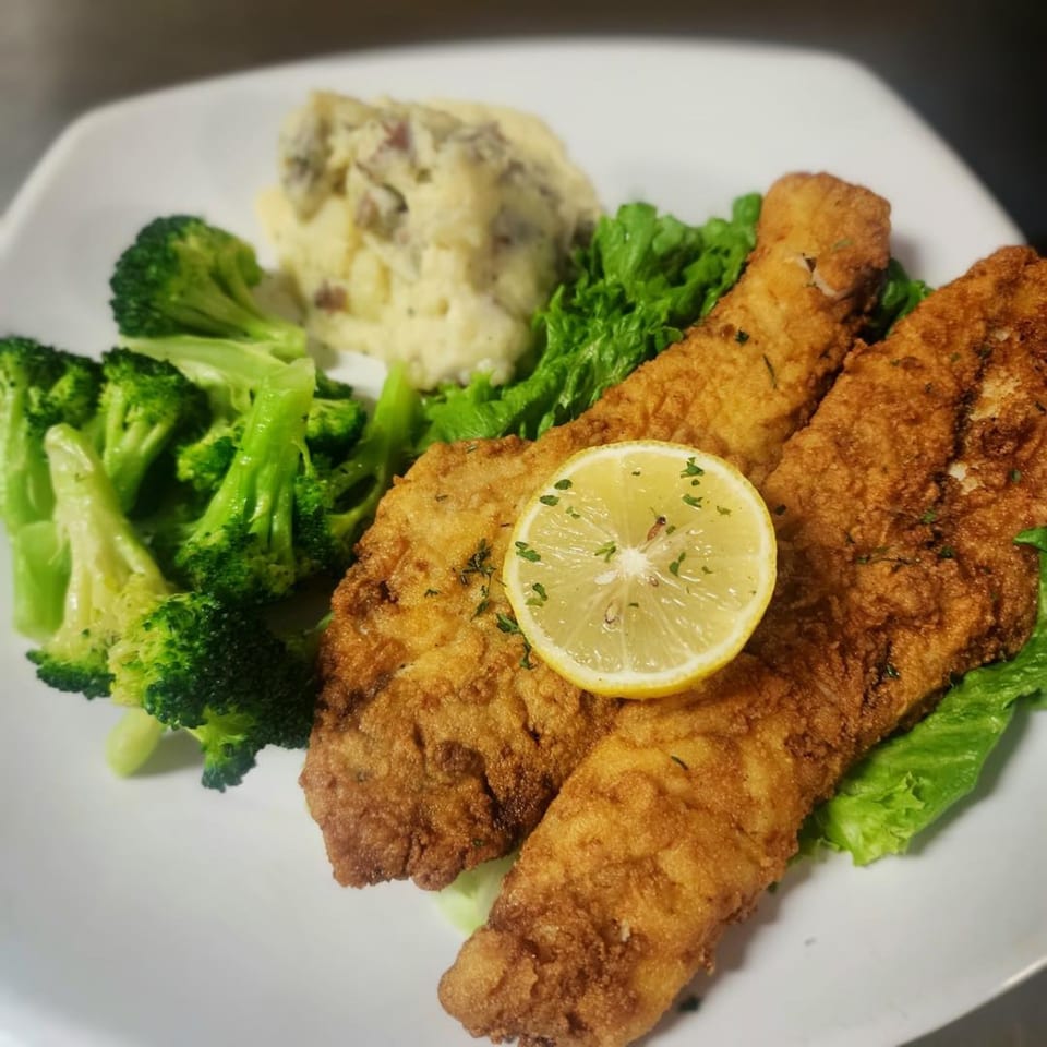 Fried fish fillets with a lemon slice, mashed potatoes, and steamed broccoli on a white plate.