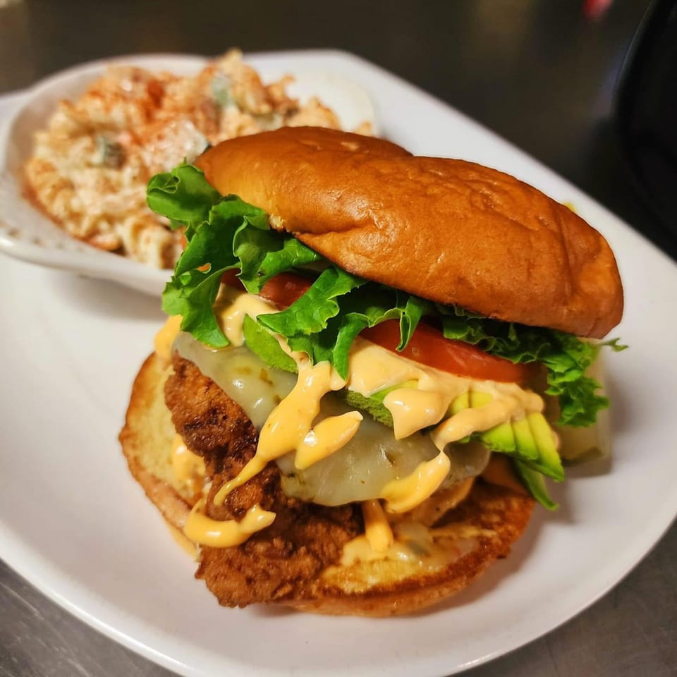 A crispy chicken sandwich with melted cheese, avocado, and sauce, served with a side of macaroni salad.
